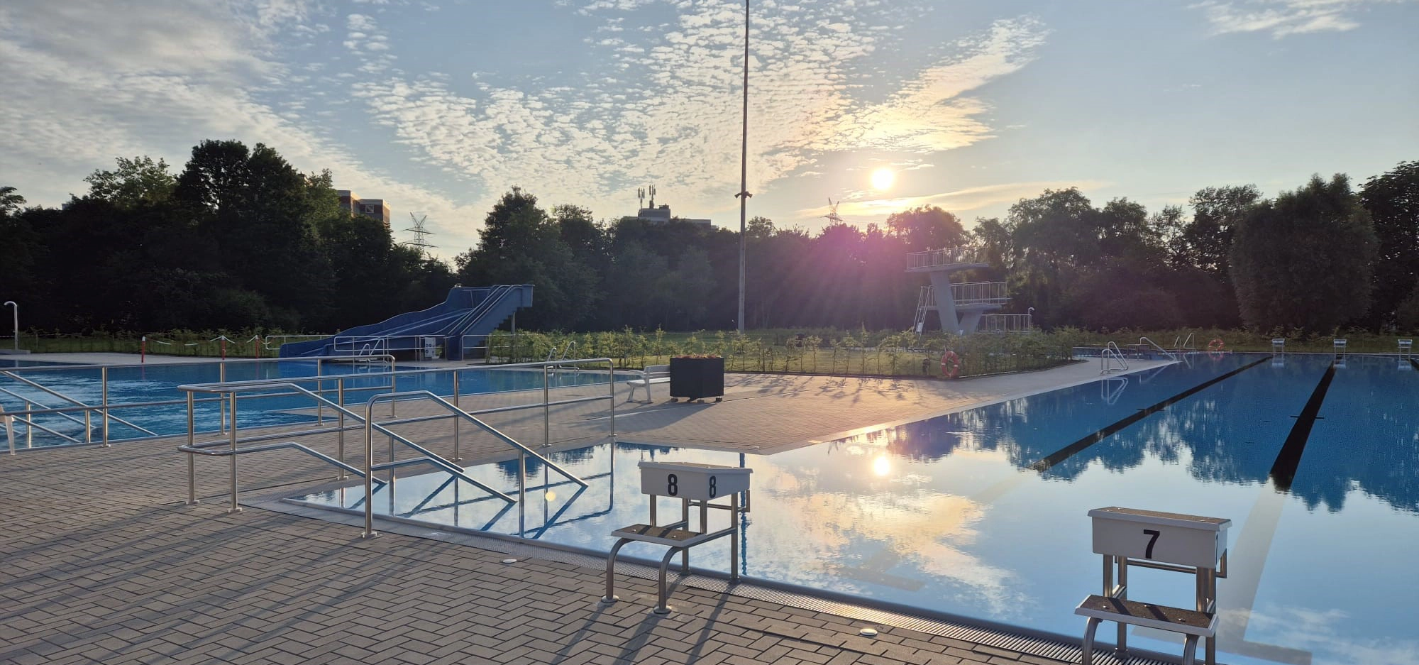 Blick auf beide Schwimmbecken mit Rutsche und Sprungturm im Hintergrund.