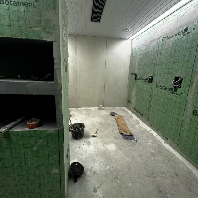 Blick in den Duschraum. Eine Wand ist gefliest. Die andere Wand und der Raumtrenner mit Ablagefächern sind mit grüner Folie verkleidet.