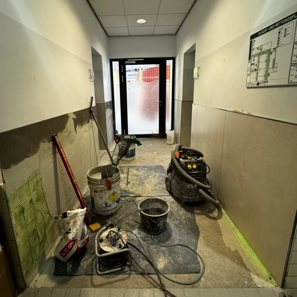 Auf einer Folie stehen Eimer und Arbeitsgeräte. Auf einer Seite sind Wandfliesen sind grün grundiert. Auf der der anderen Seite sind die neuen Fliesen an der Wand