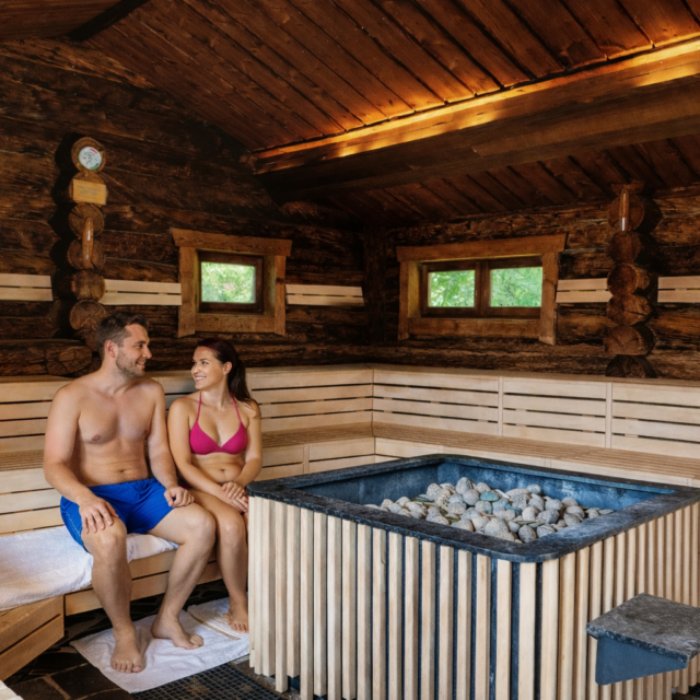 Eine Frau und ein Mann sitzen in der Eventsauna auf einer Bank. Die Frau trägt einen Bikini, der Mann trägt eine Badehose. Das Bild ist KI-generiert.