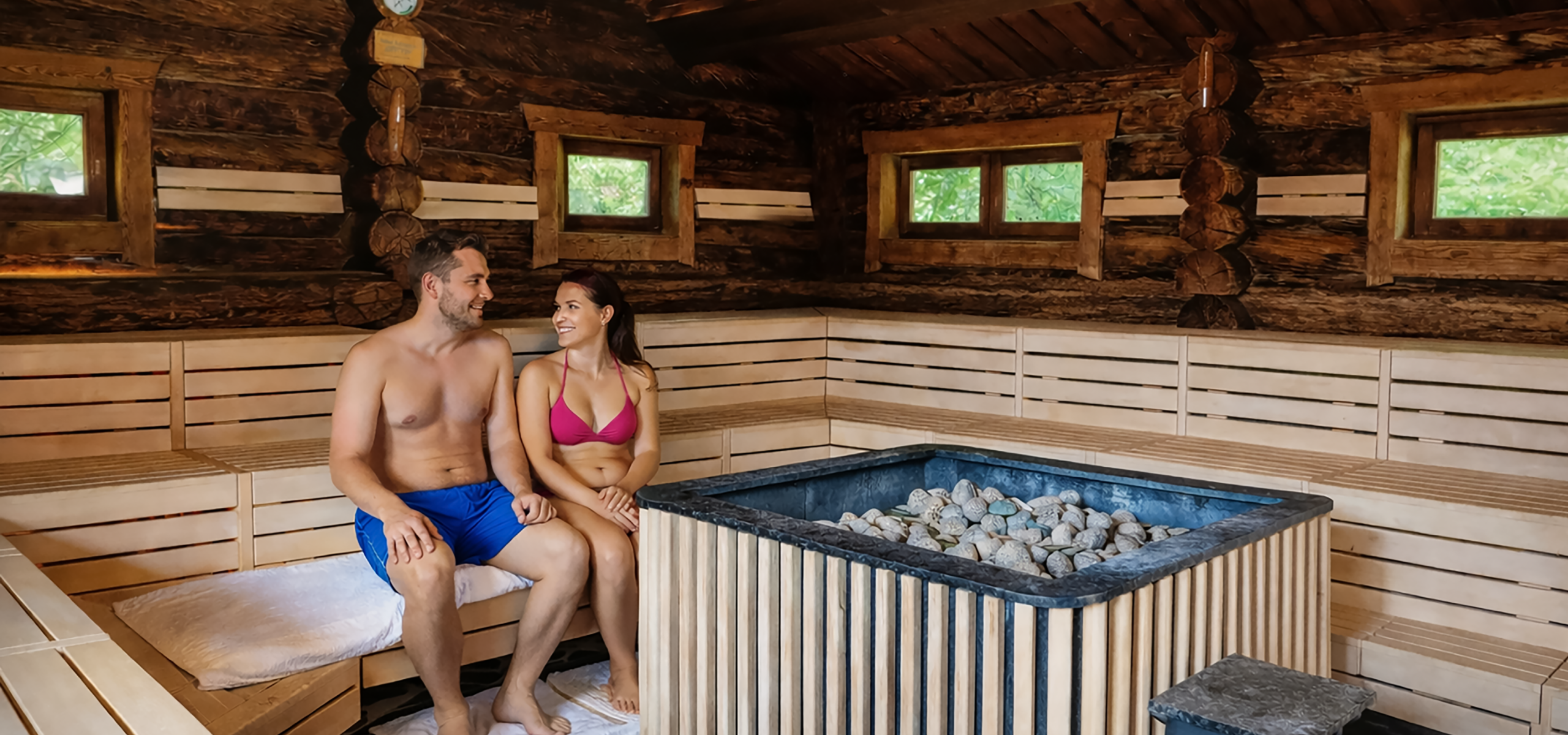 Eine Frau und ein Mann sitzen in der Eventsauna auf einer Bank. Die Frau trägt einen Bikini, der Mann trägt eine Badehose. Das Bild ist KI-generiert.